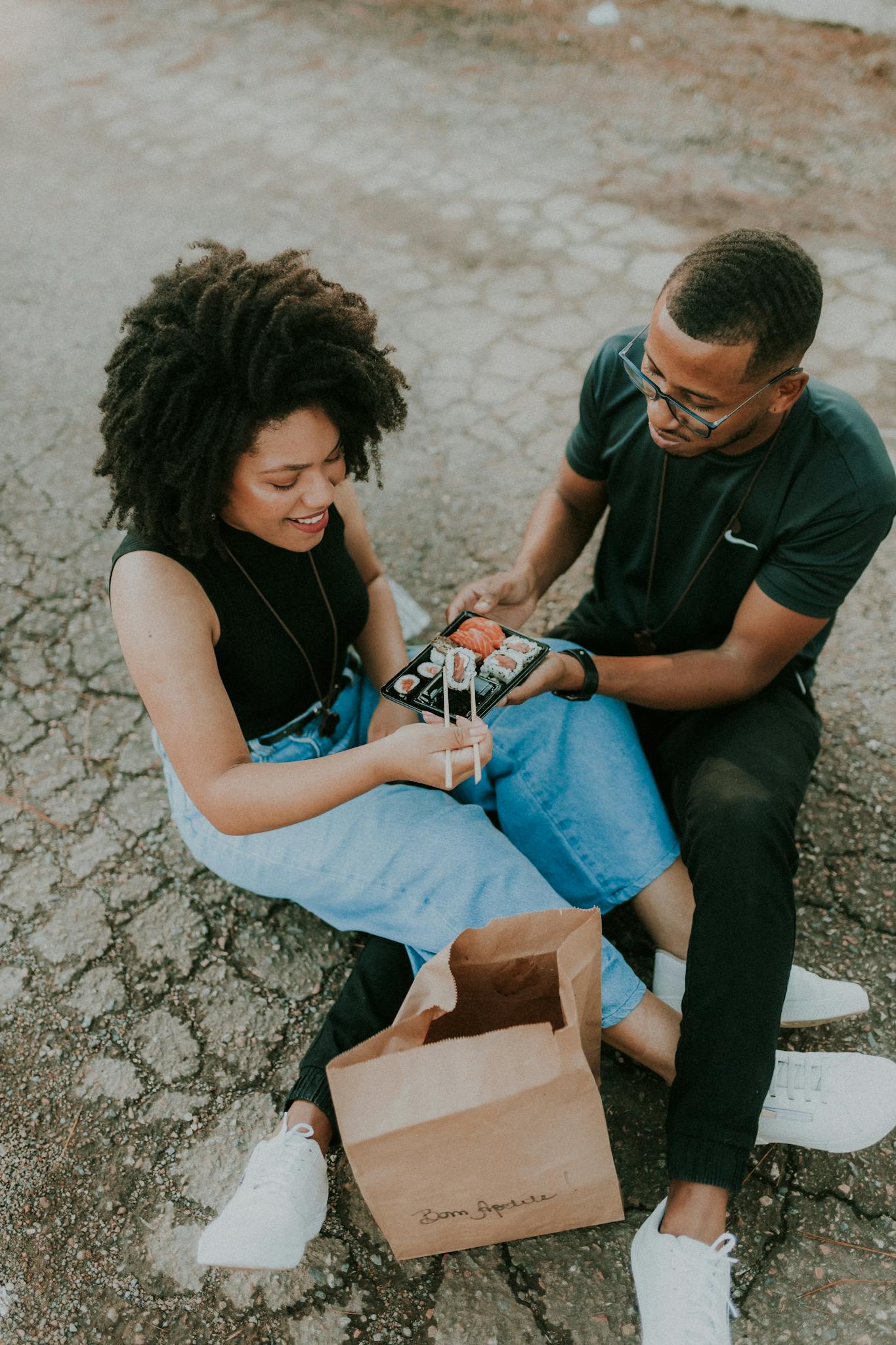 Happy couple sitting on the pavement enjoying sushi from a paper bag outdoors.
