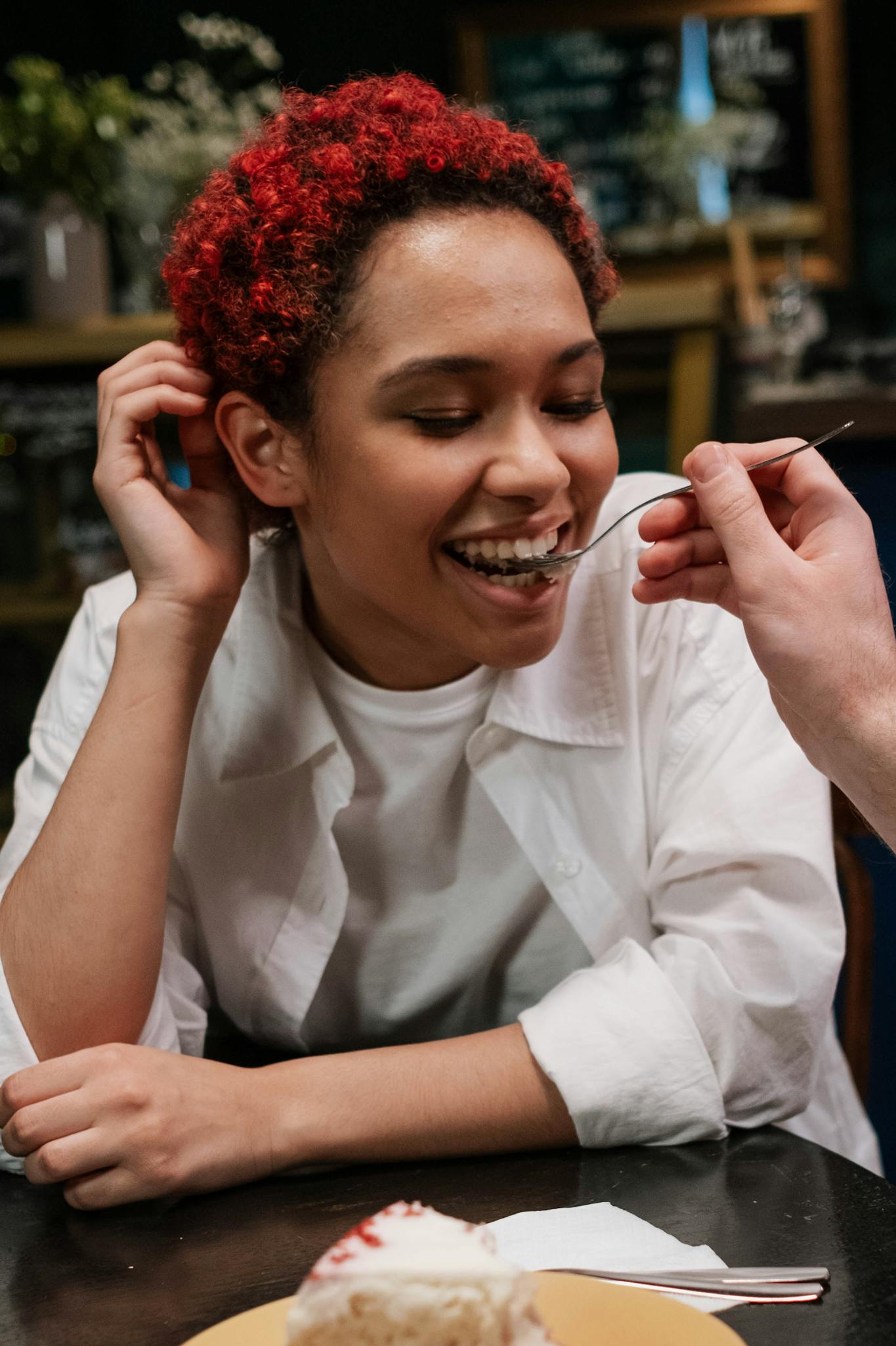 Smiling woman with red hair being fed dessert indoors, spreading happiness.